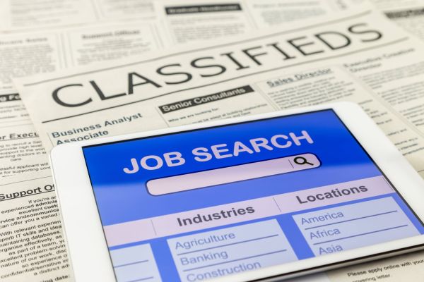 A white tablet open to an online job search board page, resting atop a stack of newspapers open to the “classifieds” section.