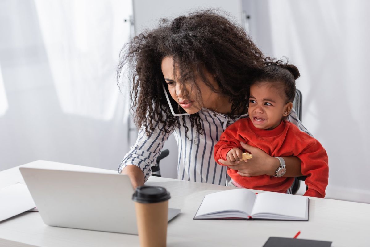 An individual seated at a table, working on a laptop with one hand while gripping a cellphone between their shoulder and cheek, holding a crying child on their lap with the other hand.