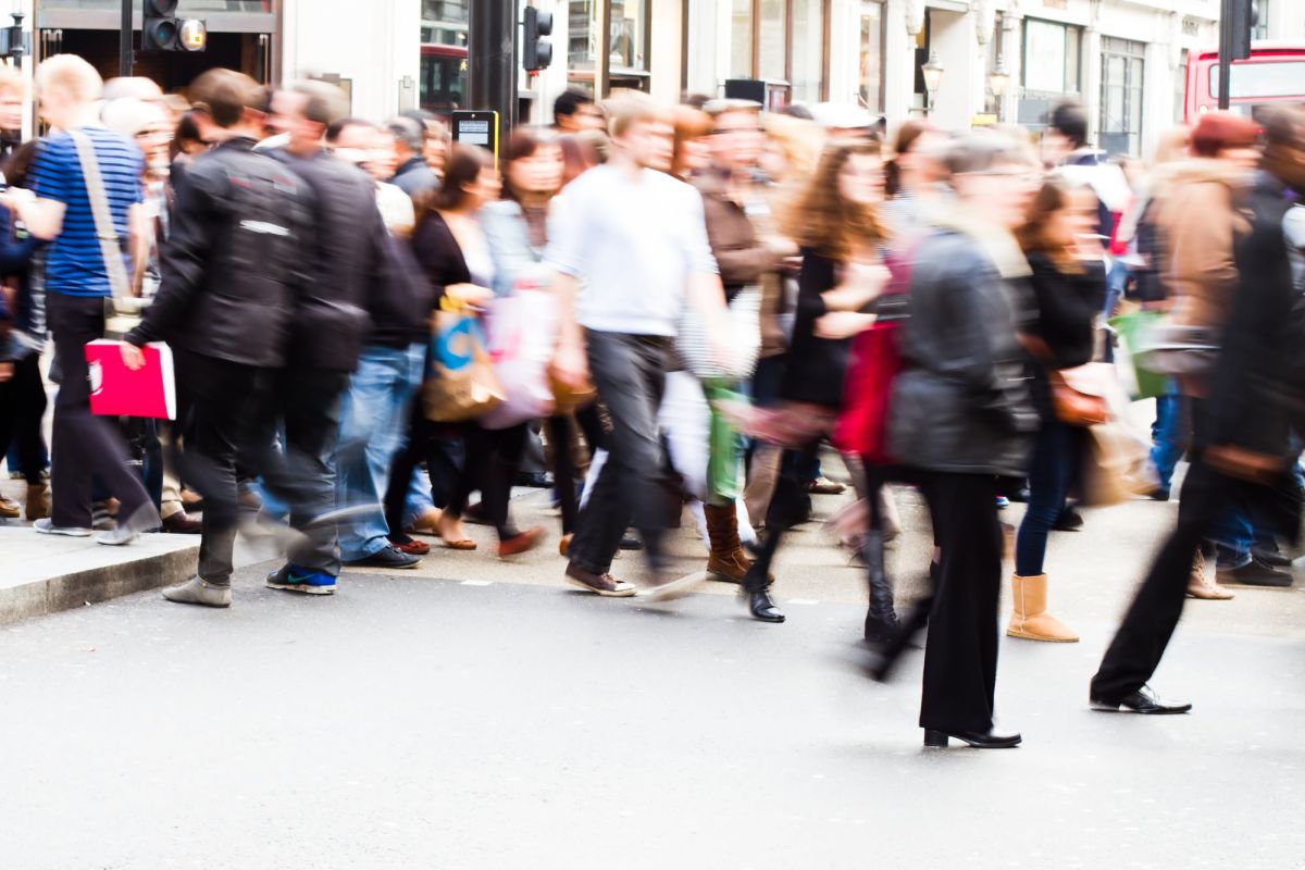 Motion-blurred image of a crowd of people moving across a crosswalk and along the nearby sidewalk of a busy city street.
