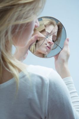 Over-the-shoulder view of an individual looking into a shattered hand mirror and closing their eyes.